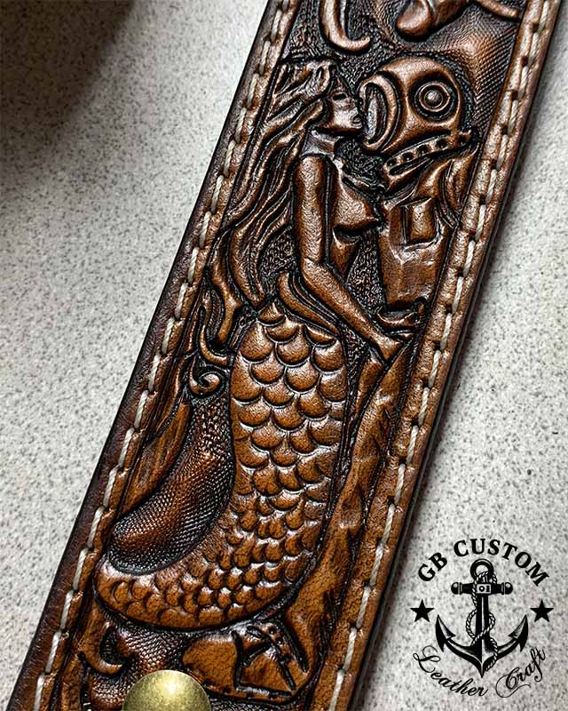 A mermaid tooled in relief on a leather belt, the scaled tail curling down the strap, reaching up to embrace a hard-hat diver in a Mark V helmet.