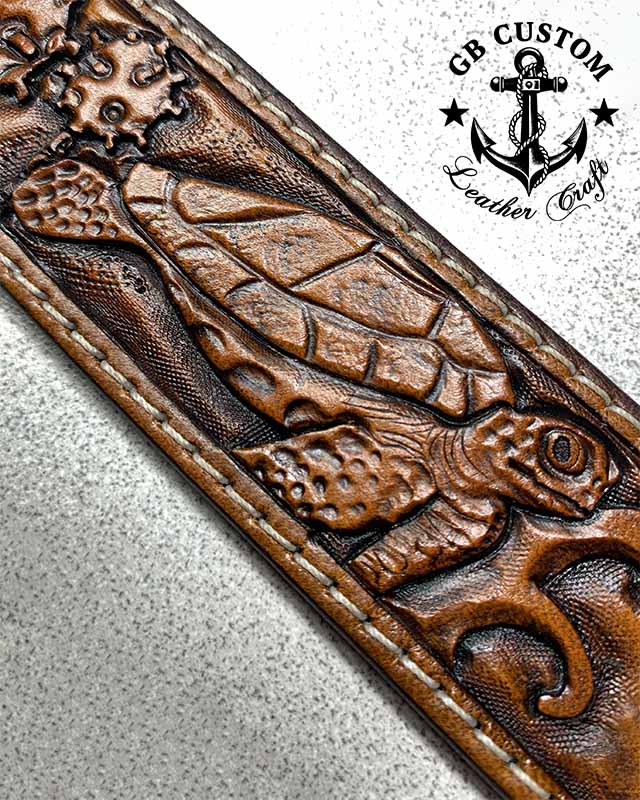 A sea turtle tooled in relief on a tan leather belt, shell and flippers textured scale by scale, with cream stitching running along the edge.