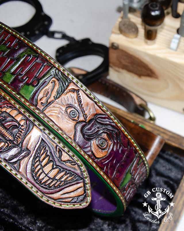 A hand-dyed belt tooled with wide grinning faces in green, purple and red, edged in green thread, resting on a workbench beside a set of handcuffs.