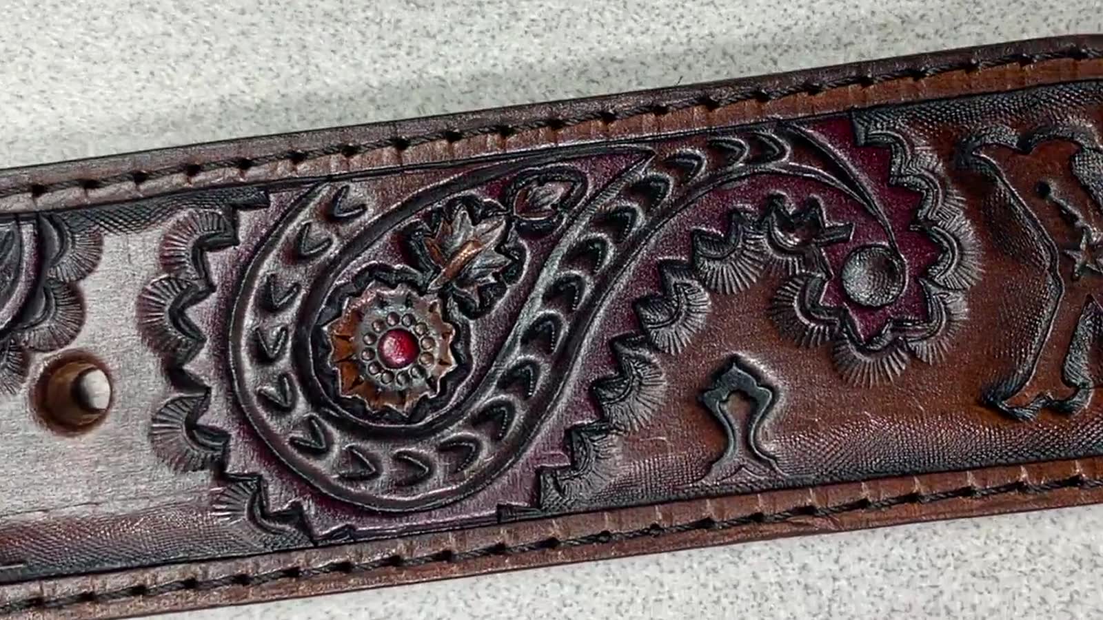 A close-up of a hand-tooled leather belt: a deep paisley scroll with layered floral rosettes, cut and shaded by hand, finished dark brown with burgundy showing in the recesses and stitched along both edges.