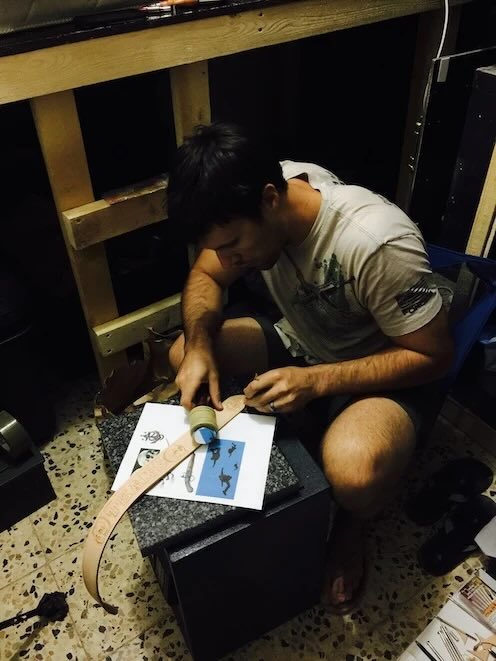 The craftsman kneeling on a workshop floor, tooling a belt blank on a granite slab with a reference drawing taped down beside it.
