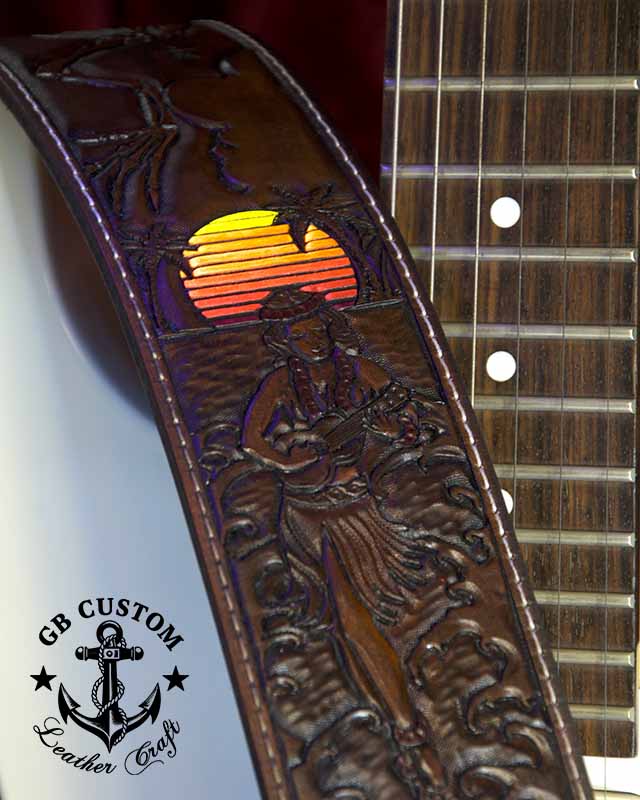 A leather guitar strap lying across a fretboard, tooled with a hula dancer playing a ukulele before a banded sunset, palm trees and breaking waves.